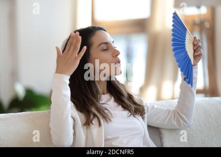 Gestresste Frau sitzt auf der Couch winkt sich mit Handventilator Stockfoto