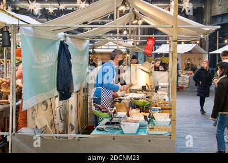 Mann mit Gesichtsmaske serviert türkische Süßigkeiten und Wraps auf einem Canopy Food Market in Coal Drops Yard, Kings Cross, London. Stockfoto