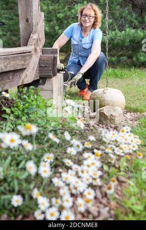 Frau mittleren Alters sitzt mit Handschuhen in der Nähe von Holzveranda und Kümmert sich um Kamillen, die im Hinterhof wachsen Stockfoto