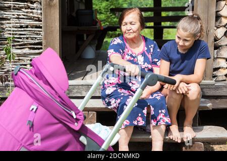 Ältere Großmutter schwingt einen Kinderwagen mit Baby, Teenager-Alter Mädchen sitzt in der Nähe, kaukasischen Menschen sind auf der Veranda in der Sommersaison Stockfoto