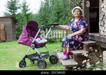 Eine Frau im Rentenalter geht mit einem Kinderwagen im Hof eines Sommerhauses, in der Nähe des Holzhauses, Hof Stockfoto