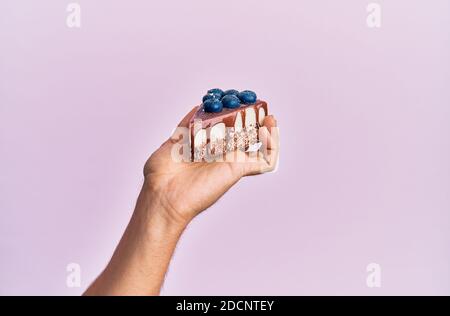 Hand des hispanischen Mannes hält Scheibe Schokoladenkuchen über isolierten rosa Hintergrund. Stockfoto