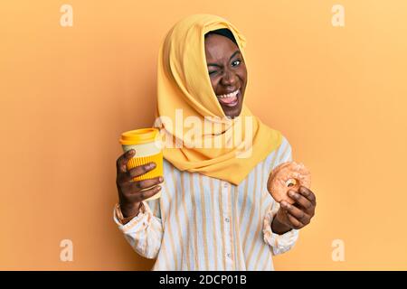 Schöne schwarze Frau trägt Hijab essen Donut und trinken Kaffee wütend und verrückt schreien frustriert und wütend, schreien vor Wut. Wut und Stockfoto