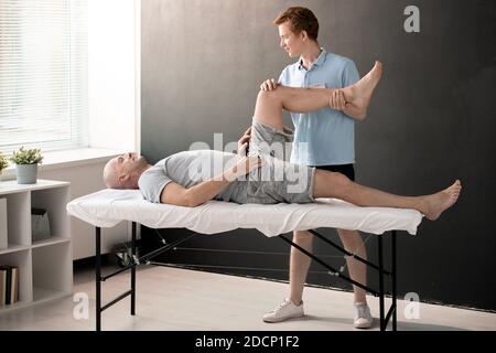 Männlicher Physiotherapeut, der das Bein des Patienten im Knie gebeugt hält und dabei hilft Er Stockfoto