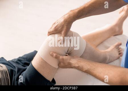 Hände des männlichen Klinikpersonals wickeln Knie des Patienten mit Flexible Bandage Stockfoto