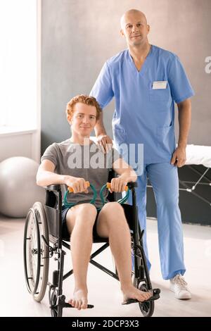 Reifer Arzt in blauer Uniform steht bei jungen männlichen Patienten Im Rollstuhl Stockfoto