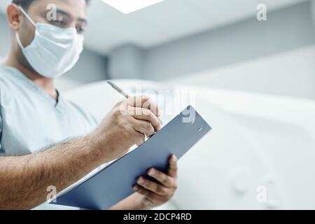 Hände von reifen Mischrasse männlichen Arzt in Uniform und Maske Notizen machen Stockfoto