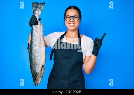 Junge schöne asiatische Mädchen Fischhändler Verkauf frische rohe Lachs lächelnd Glücklich zeigen mit Hand und Finger zur Seite Stockfoto