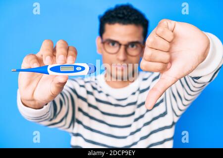 Junge gutaussehende hispanische Mann hält Thermometer mit wütenden Gesicht, negative Zeichen zeigt Abneigung mit Daumen nach unten, Ablehnung Konzept Stockfoto