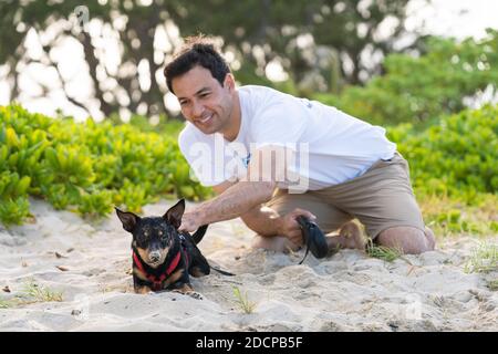 Der junge Mann kniet sich nieder, um einen schwarzen Hund mit Sand zu streicheln Am Strand Stockfoto