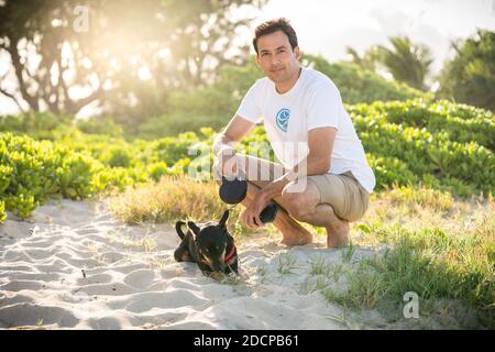 Junge fit männlichen Kniebeugen am Strand mit seinem kleinen Schwarzer Hund auf Hawaii Stockfoto