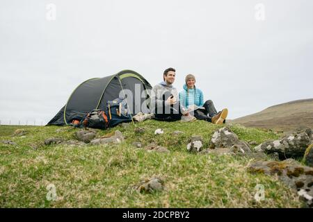 Fröhlicher Mann und Frau mit Buch sitzen in der Nähe des Zeltes gegen Grauer Himmel in hügeligem Gelände Stockfoto