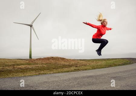 Frau springt mit ausgestreckten Armen während des Trainings gegen Windmühle in Natur Stockfoto