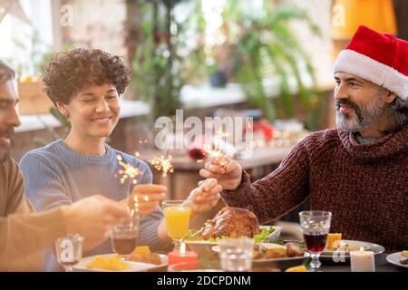 Zwei glückliche Männer und eine junge Frau halten bengalische Lichter über Festlicher Tisch serviert Stockfoto