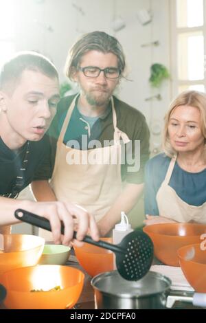 Junger männlicher Koch-Coach und seine beiden Auszubildenden in Schürzen Biegen über den Arbeitsplatz Stockfoto