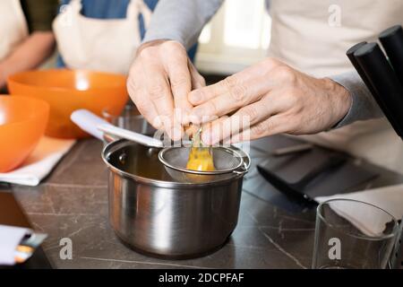 Die Hände des älteren Mannes in der Schürze zerbrechen Ei in Sieb Über Metallpfanne für Tisch Stockfoto