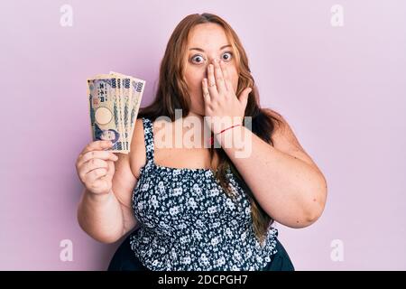 Plus Größe kaukasische junge Frau hält japanische Yen Banknoten decken Mund mit Hand, schockiert und Angst vor Fehler. Überrascht Ausdruck Stockfoto