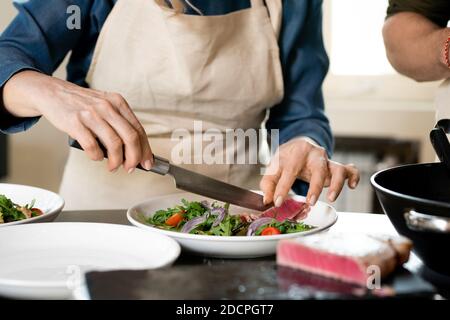 Hände von reifen weiblichen Putting Scheibe geräuchertes Fleisch auf Teller mit Salat Stockfoto
