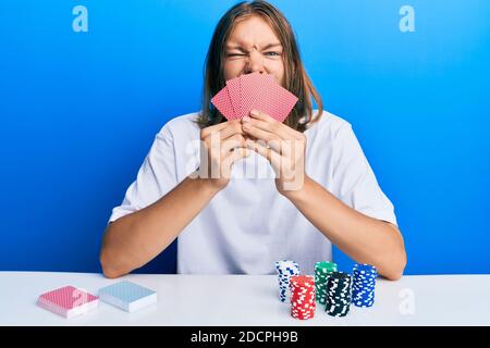 Schöner kaukasischer Mann mit langen Haaren spielen Glücksspiel Poker Abdeckung Gesicht mit Karten aufragenden Zunge aus glücklich mit lustigen Ausdruck. Stockfoto