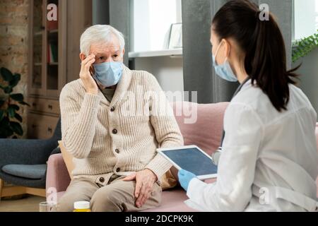 Kranker älterer Mann in schützender medizinischer Maske, der sich beim Arzt beschwert Über Kopfschmerzen Stockfoto