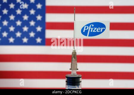 Stafford / Vereinigtes Königreich - November 22 2020: Pfizer-Impfstoff Covid-19 Konzept. Spritzennadel und Pfizer Aufkleber darauf, verschwommene Flagge der United Sta Stockfoto