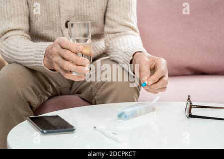 Hände von älteren Mann in Casualwear halten Pille und Glas Von Wasser über dem Tisch Stockfoto