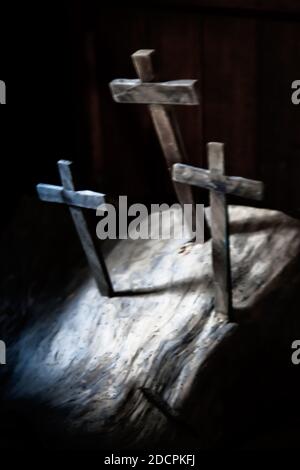 Eine verschwommene Ansicht von drei hölzernen Kreuzen, die in einen eingestochen wurden Platte in einem dreifaltigkeitsmuster in einem dunklen Innenraum mit Licht strahlend nach unten für spirituelle Konzept Stockfoto