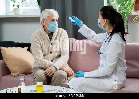Junge Ärztin in Whitecoat Messung der Körpertemperatur von Kranken Älterer Herr Stockfoto