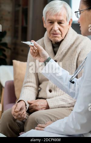 Älterer Mann sitzt auf der Couch neben einer jungen Ärztin Mit Thermometer Stockfoto
