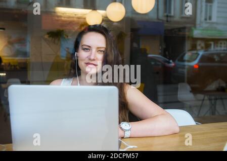 Blick durch das Fenster mit der Stadt Spiegelung der lächelnden Geschäftsfrau im Headset arbeiten auf Laptop sitzen im Café, Coworking. Remote-Arbeit, soziale Netzwerke Stockfoto