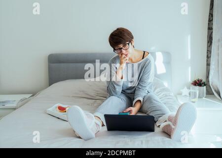 Verwirrt und nachdenklich kaukasische Frau in Brille und lässige Hauskleidung mit einem Laptop sitzen auf ihrem Bett und Frühstück. Remote-Arbeiten von Stockfoto