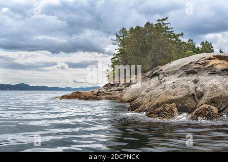 An einem unruhigen Tag ragt ein felsiger Punkt in das gekräuselte Wasser, während drohende Wolken über den Inseln in der Ferne zu schweben scheinen. Stockfoto