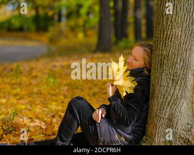 Ein schönes junges blondes Mädchen sitzt auf gefallenen Herbstblättern im Park und lehnt ihren Rücken gegen einen Baumstamm. Das Mädchen hält gelbe Ahornblätter Stockfoto