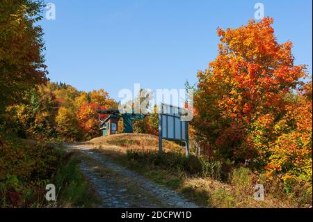 Bergstraße durch einen Mischwald in feurigen Herbstfarben. Oberes Terminal eines Sessellifts und Weginformationsschild. Mt. Mansfield, Stowe, Vermont, USA Stockfoto