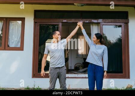Portrait von jungen erwachsenen asiatischen Paar hält Hände zusammen mit Zuhause Interieur im Hintergrund. 30s glücklich reifen Mann und Frau lächeln. Immobilien- und Immobilienleistungskonzept Stockfoto