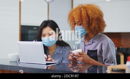 Zwei junge Frauen asiatisch und schwarz mit Gesichtsmasken Browsen Social Media auf dem Laptop. Hochwertige Fotos Stockfoto