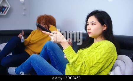 Gelangweilte Rommates, Studenten sitzen auf der Couch und schauen sich das Fernsehen an, das sich über das Geschwätz und den Verzehr von Pralinen unterhalten. Hochwertige Fotos Stockfoto