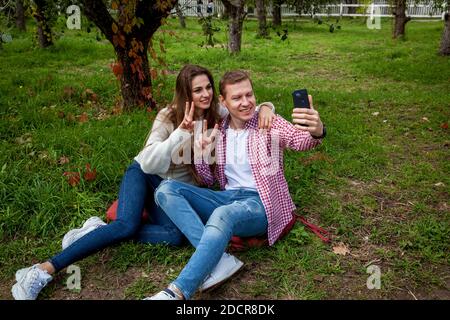 Ein junges Paar verbringt Zeit zusammen im Park, sitzt auf dem Gras, macht Selfies am Telefon. Der Typ mit einem Mädchen mit Gadgets im Park Stockfoto