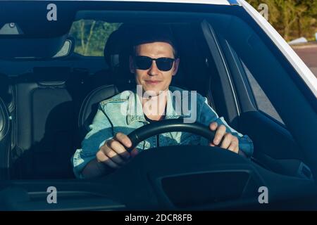Kerl in Jeansjacke und schwarze Brille lächelt an der Rad eines Autos Stockfoto