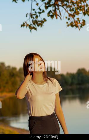 Schöne junge Brünette Frau auf dem Flussufer bei Sonnenuntergang. Weichzeichner, Unschärfe. Stockfoto