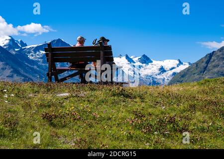 Zwei Wanderer sitzen auf einer Bank bei Muottas Muragl, den schneebedeckten Bergen des Oberengadins in der Ferne. Stockfoto