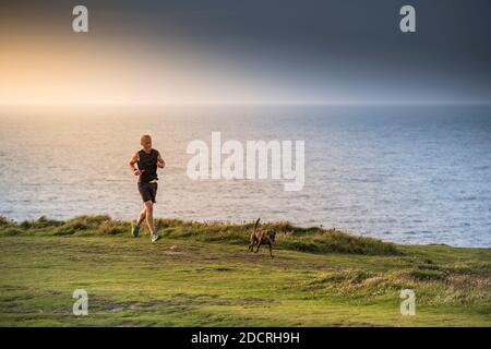 Ein Mann, der mit seinem Hund auf dem Pentire Point East in Newquay in Cornwall einen Fußweg entlang läuft. Stockfoto