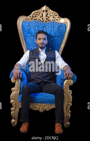 Junger bärtiger Mann in formeller Weste sitzt auf einem blauen Maharaja Stuhl. Schwarzer Hintergrund. Stockfoto
