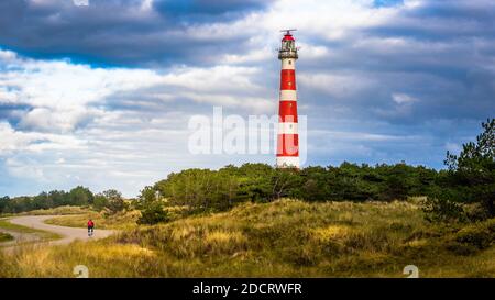 Ein rot-weißer Leuchtturm in den Dünen an einem bewölkten Tag mit einer Straße, auf der ein Radfahrer in einem roten Hemd radelt. Stockfoto