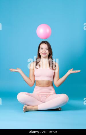 Eine lächelnde Frau in rosa Sportkleidung sitzt mit gekreuzten Beinen und wirft einen Ball, die Arme ausgestreckt, isoliert auf einem blauen Hintergrund Stockfoto