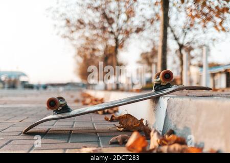 Skateboarding. Ein umgedrehtes Skateboard liegt auf dem Bürgersteig, mit heruntergefallenen Blättern. Im Hintergrund eine leere Straße der Stadt. Nahaufnahme. Stockfoto