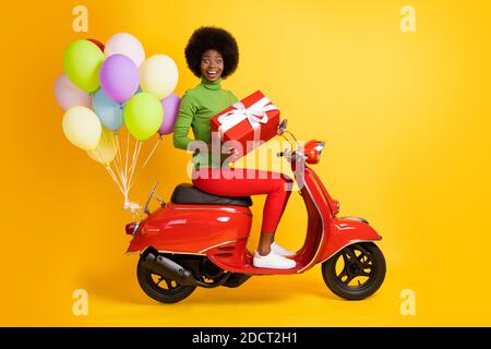 Foto-Porträt von Brünette afroamerikanische Frau mit Geschenkbox Auf dem Motorrad mit Luftballons tragen lässige rote und grüne Kleidung Isoliert auf lebhaft Stockfoto