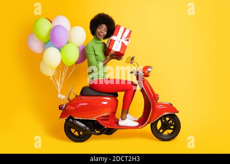 Foto-Porträt von brünett afroamerikanischen Mädchen halten rot gewickelt Geschenk Reiten Fahrrad mit Luftballons isoliert auf lebendige gelb Farbiger Hintergrund Stockfoto