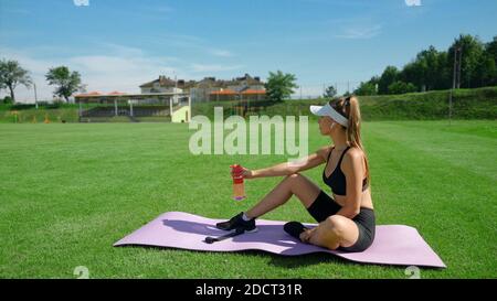 Seitenansicht der jungen fittigen Frau, die Flasche mit Wasser hält, auf der Matte sitzend, Stadionfeld im Sommer sonnigen Tag. Sportliche Mädchen tragen Sport-Outfit mit Ruhe auf grünem Gras. Konzept des Sports, Workout. Stockfoto