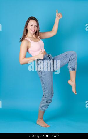 Eine ganze Länge lachende Frau in Jeans und einem rosa Top tanzt und gibt einen Daumen nach oben isoliert auf einem Blauer Hintergrund Stockfoto